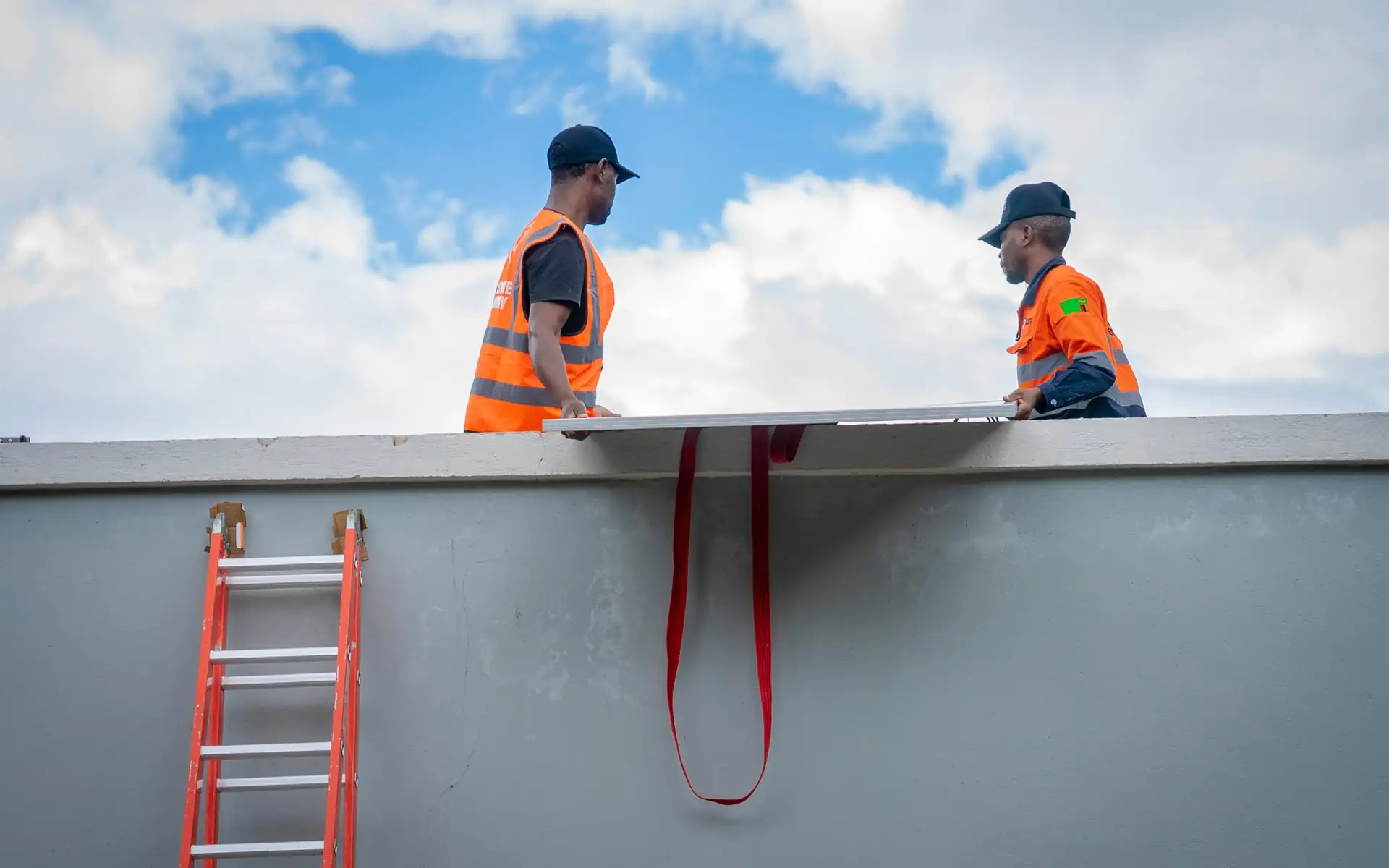 Two EcoHive installers on a roof edge lifting a solar panel against a blue sky